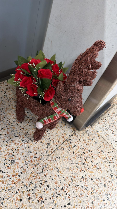 Wicker Reindeer - A rustic handcrafted reindeer decoration made from natural woven materials, finished with a festive scarf and a vibrant arrangement of red flowers and greenery.